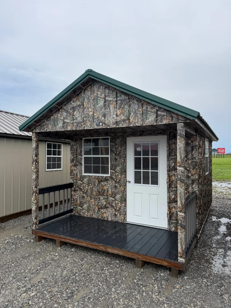 Camouflage-style portable cabin with green metal roof, covered front porch, and white entry door