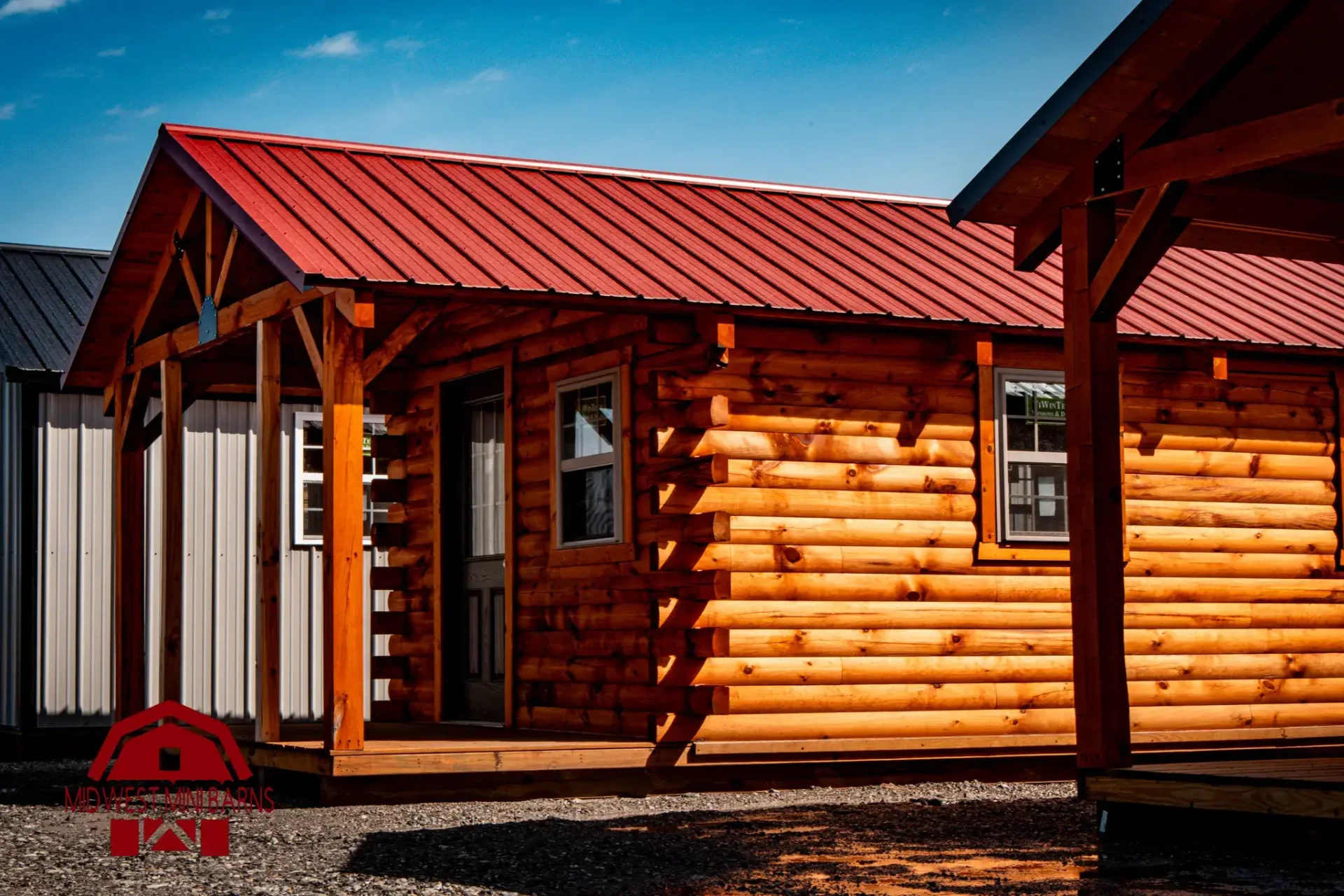 Log cabin with a red metal roof and covered porch on a Midwest Mini Barns lot