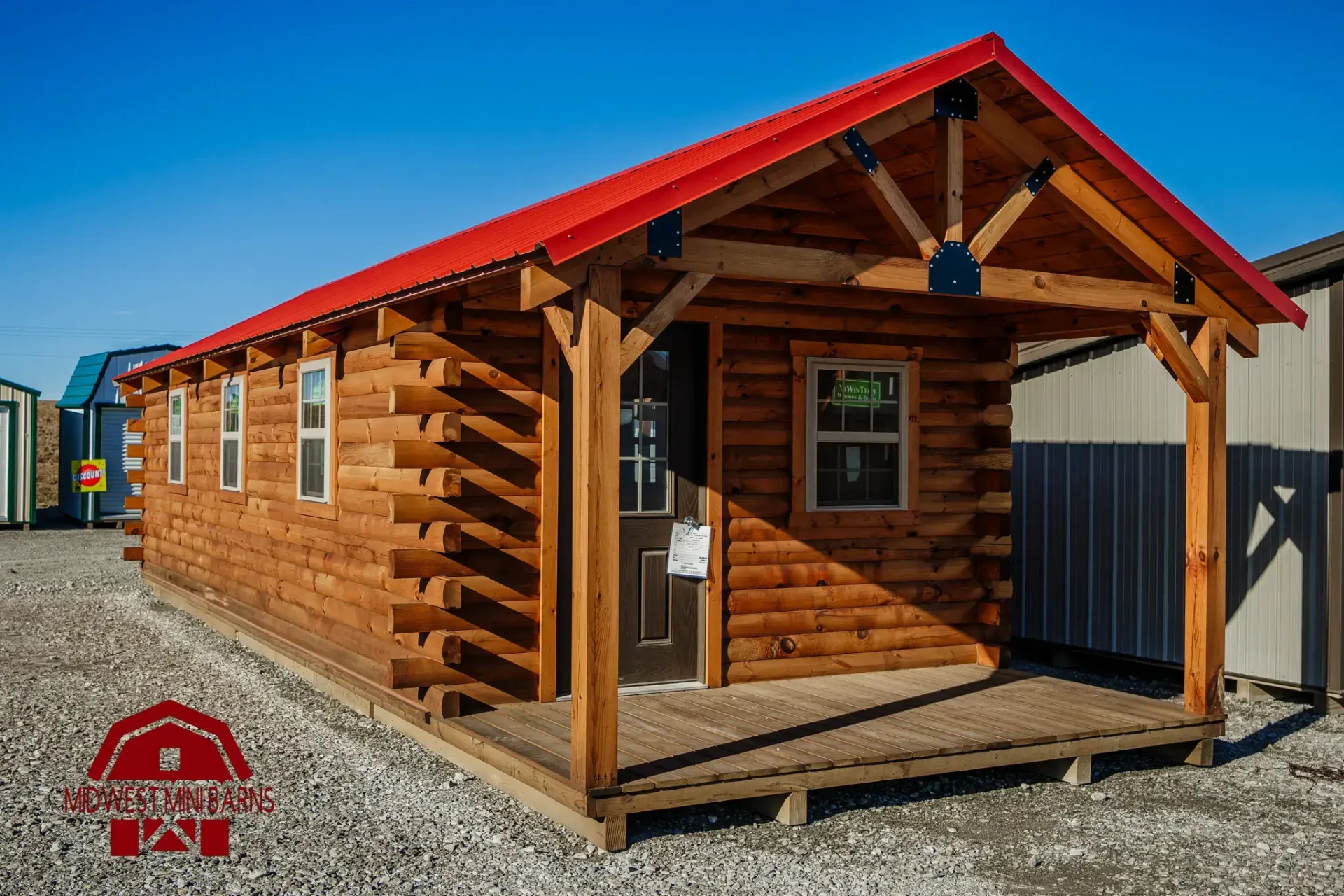 Log cabin shell with a red metal roof and covered porch on a display lot
